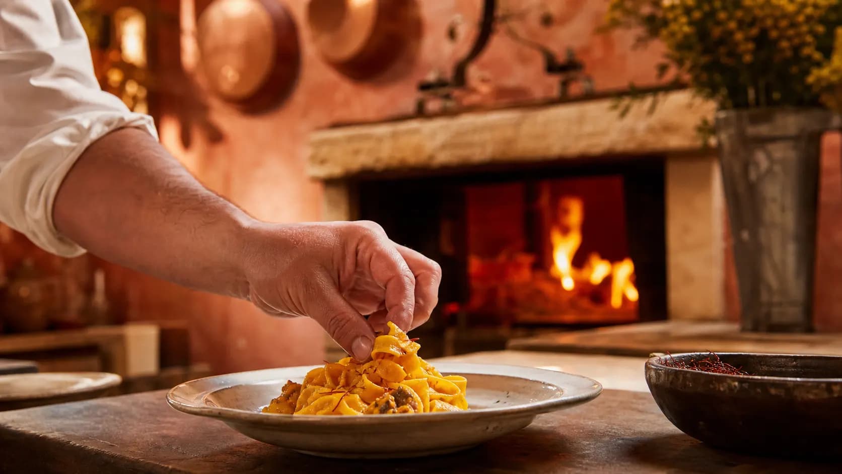 Chef plating saffron pasta beside a wood-fired hearth